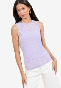 Top senza maniche in pizzo lavanda con motivi floreali e una texture trasparente, indossato con pantaloni bianchi, che mette in risalto il design intricatamente dettagliato del capo.