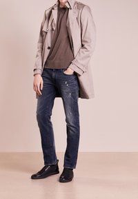 Abrigo trench en beige claro, suéter marrón, jeans oscurecidos de mezclilla azul y zapatillas negras texturizadas con acentos brillantes.