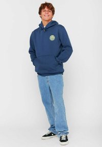 Felpa blu navy con tasca frontale e coulisse, caratterizzata da un logo verde rotondo. Indossata con jeans azzurri chiari e scarpe nere.