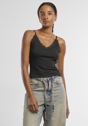Femme aux cheveux tressés portant un débardeur noir bordé de dentelle et un jean clair avec une ceinture en tissu nouée, debout devant un fond uni.