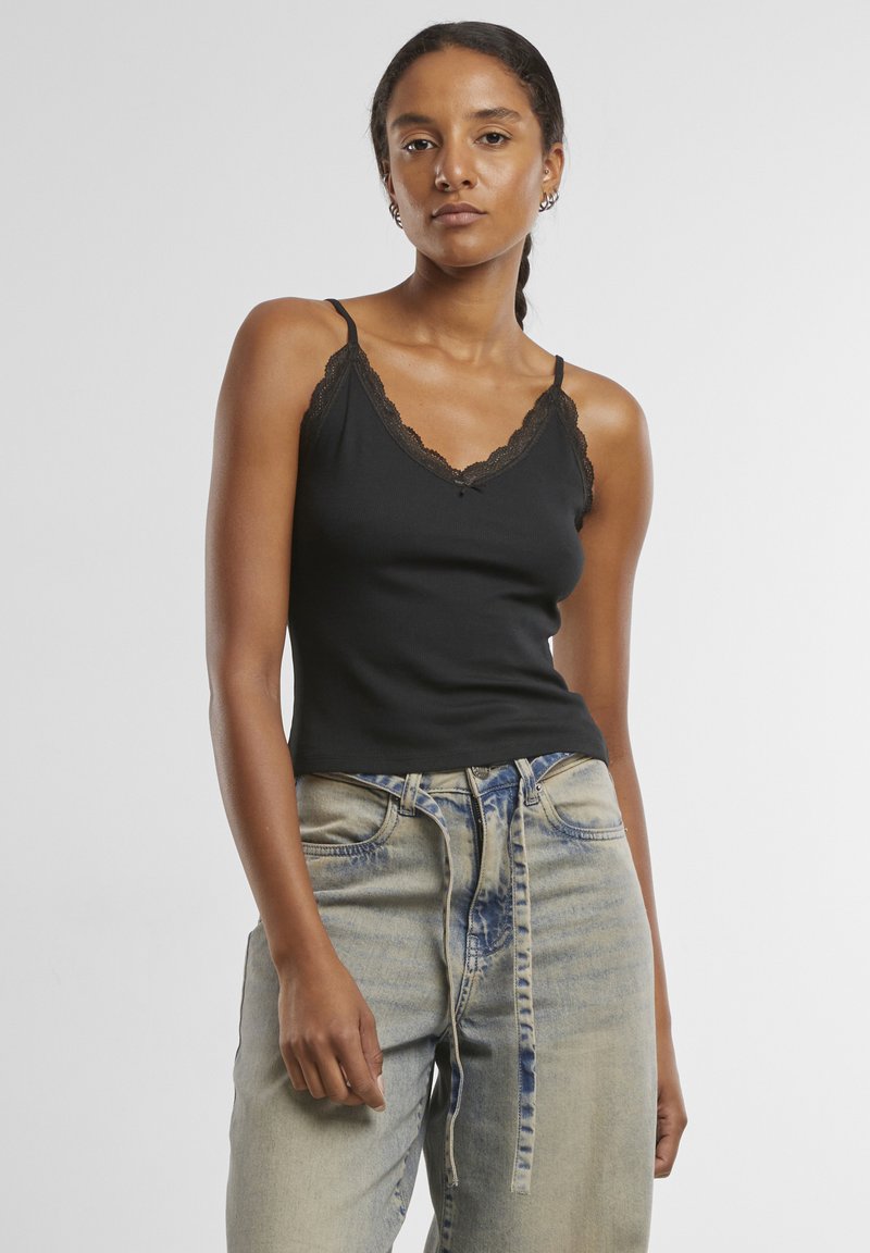 Femme aux cheveux tressés portant un débardeur noir bordé de dentelle et un jean clair avec une ceinture en tissu nouée, debout devant un fond uni.