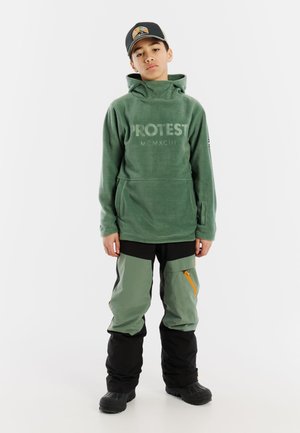 Grüne Fleece-Hoodie mit einer Kängurutasche und Logo, kombiniert mit schwarzen und grünen Snowboardhosen mit orangefarbenem Reißverschluss und lockerer Passform.