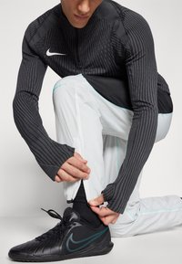 Sportliches Outfit mit einem schwarzen Langarmoberteil mit gerippter Struktur und einer weißen Hose mit Reißverschlussbündchen. Schwarze Sneakers mit türkisfarbenen Akzenten.