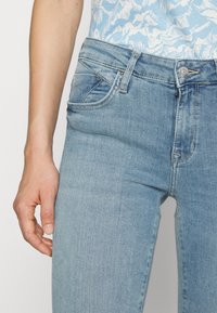 Jeans de denim azul claro con un diseño ajustado, con bolsillos delanteros y un cierre estándar de botón. La tela tiene una textura suave.