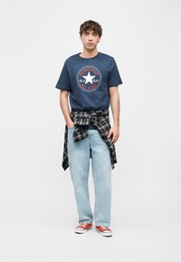 Νεαρός άνδρας που φοράει μπλε μπλουζάκι Converse All Star, ανοιχτόχρωμο τζιν, μαύρο καρό πουκάμισο δεμένο στη μέση και κόκκινα αθλητικά παπούτσια Converse, στέκεται όρθιος.