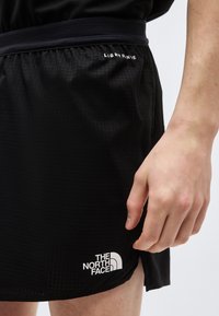 Nærbilde av en person som har på seg svarte shorts med logoene "The North Face" og "Light Rinse" på stoffet nær livet og kanten.