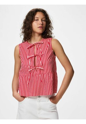 Mladá žena s kučeravými vlasmi nosí červenobiely pásikavý tielko bez rukávov s tromi mašličkami vpredu, doplnené bielymi nohavicami, ruky v vreckách.