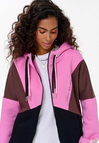 Kurtka z kapturem w kolorowe bloki, z różowymi, brązowymi i czarnymi panelami, z trokami i zamek błyskawiczny; wykonana z miękkiego materiału.