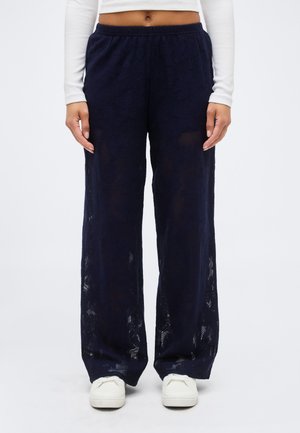 Femme portant un pantalon large en dentelle bleu marine, un chemisier blanc à manches longues et court, ainsi que des baskets blanches, debout devant un fond uni.