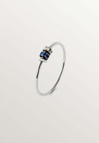 Anillo de metal plateado con una banda suave y delgada que presenta un grupo de piedras azules y transparentes en la parte superior, exhibiendo un diseño circular.