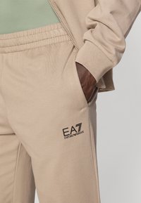 Bēša sporta bikses ar elastīgu jostu, sānu kabatām un melnu EA7 Emporio Armani logotipu uz augšstilba. Gluda tekstūra.