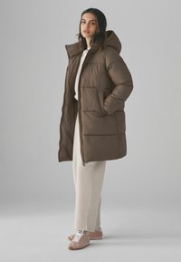 Manteau matelassé marron avec capuche, doté d'une fermeture éclair sur le devant et de poches latérales. Associé à un survêtement crème et des sneakers roses. Texture lisse.