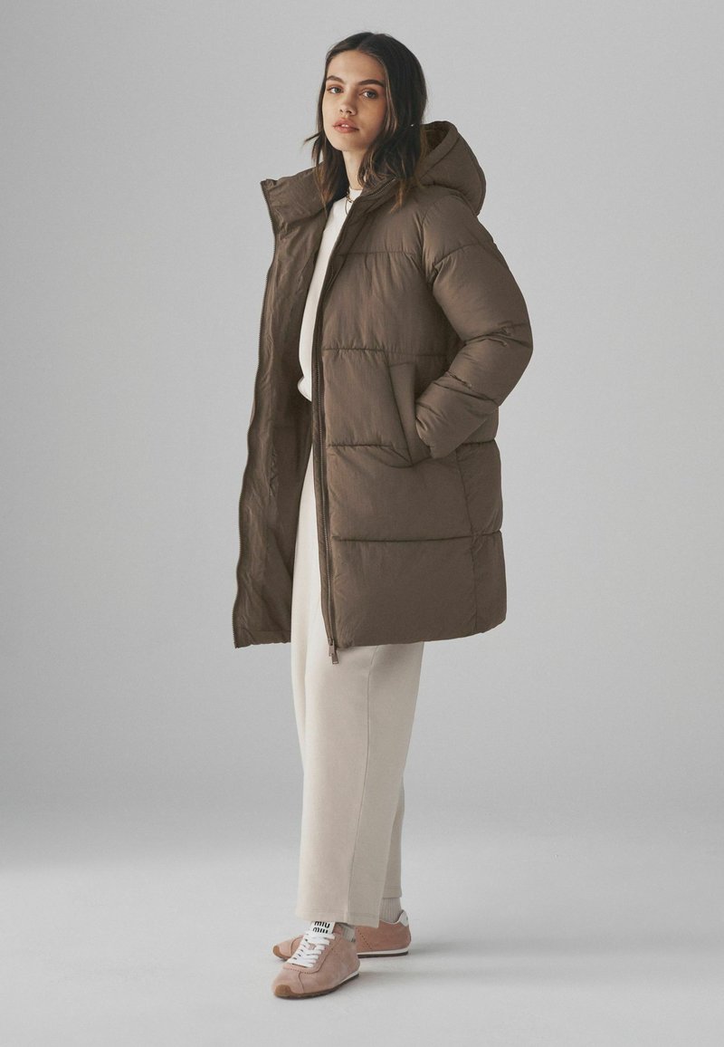 Manteau matelassé marron avec capuche, doté d'une fermeture éclair sur le devant et de poches latérales. Associé à un survêtement crème et des sneakers roses. Texture lisse.