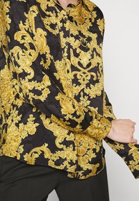Czarna i złota koszula z barokowym wzorem z długim rękawem noszona z czarnymi spodniami, ręka dostosowująca mankiet lewego rękawa.