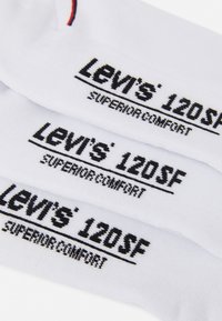 Hvide bomuldsstrømper med sort trykt tekst, der står "Levi's 120SF SUPERIOR COMFORT," med en rød accentstribe foroven.