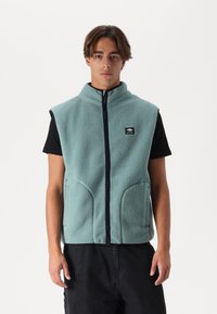 Vesta din fleece de culoare albastră deschisă, cu guler înalt, fermoar frontal și buzunare laterale. Purtată peste un tricou negru, cu un logo mic pe piept.
