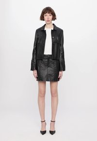 Giacca di pelle nera con dettagli di frange, abbinata a una minigonna di pelle nera con accenti di laccatura. Il modello indossa tacchi alti neri.