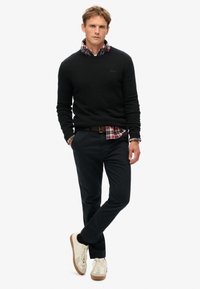 Hombre vistiendo un suéter negro sobre una camisa de cuadros rojos, pantalones oscuros y zapatillas blancas, de pie con una pierna cruzada y las manos en los bolsillos.