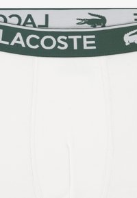 Hvide bomuldsboxershorts med en grøn elastisk linning med "LACOSTE"-branding og en krokodillelogo.