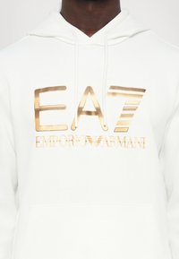 Sweatshirt branco com um grande logo metálico dourado a dizer "EA7 EMPORIO ARMANI." O tecido parece macio e possui um bolso canguru.