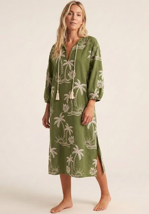 Mujer que lleva un vestido midi verde con patrones blancos de palmeras tropicales y sol, mangas largas, lazos delanteros y abertura lateral, de pie descalza.