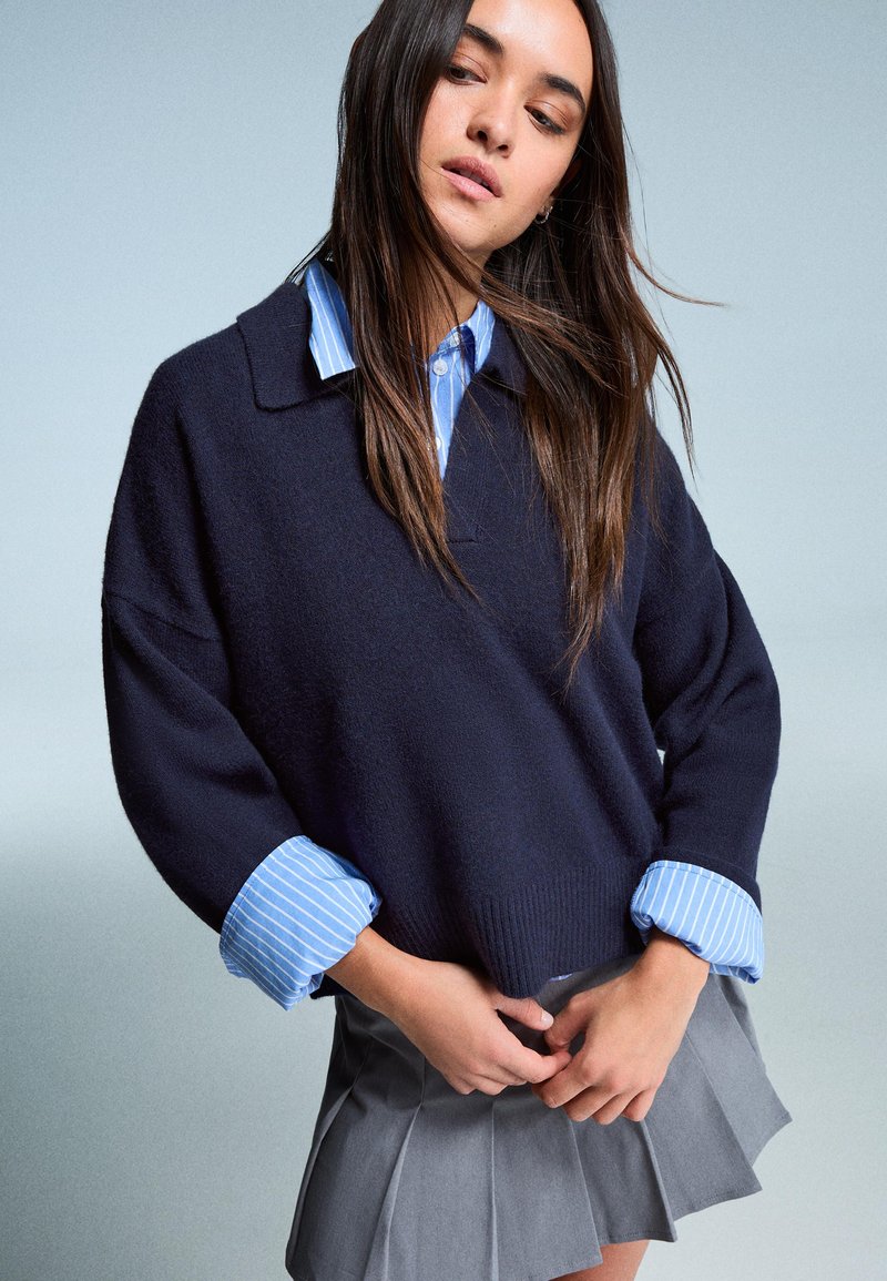 Mornarica pulover s opuštenim krojem, sa ovratnikom i prugastom plavom košuljom ispod. Sivi plisirani suknja. Glatka tekstura, ležerno slojevito.