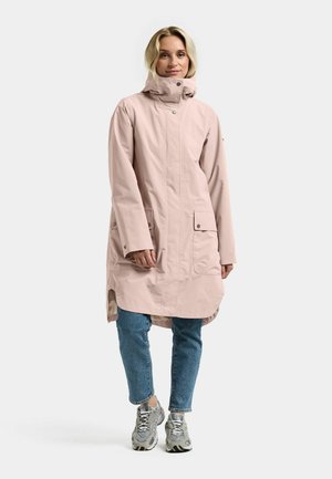 Lyserød vandtæt parka med høj krave, trykknapper, to forlommer og en længere bagkant, båret over blå jeans og sneakers.