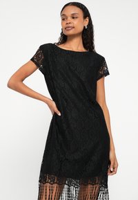 Melna mežģīņu kleita ar īsām piedurknēm, brīva piegriezuma un ar bārkstīm apakšmalā. Visu apģērbu rotā teksturēts ziedu raksts.