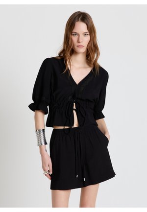 Femme portant un chemisier noir à encolure en V avec des manches bouffantes et un short ample assorti, accessoirisée d'un bracelet manchette en argent et de bagues.