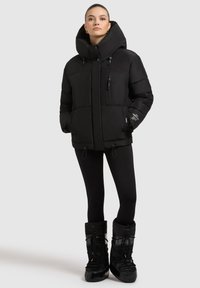 Sort polstret jakke med høj krave, lynlåslomme og snoretræk i bunden. Kombineret med sorte leggings og sorte skalstøvler.
