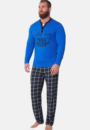 Man wearing a blue long-sleeve shirt with black text and black plaid pants standing barefoot on a white background.
