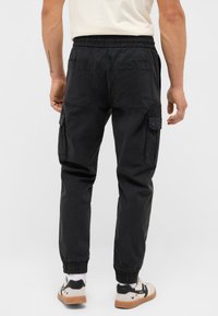 Man wearing black cargo jogger pants and white sneakers, standing with back facing the camera on plain light background.