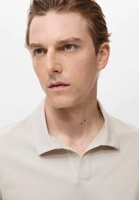 Ljusbeige polo med texturerat tyg, korta ärmar och klassisk krage. Modellen har ett neutralt uttryck och ljusbrunt hår.