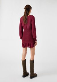 Vestido burdeos con mangas largas y cintura fruncida, con un dobladillo volante, combinado con botas altas de color verde oliva, situado frente a un fondo neutro.