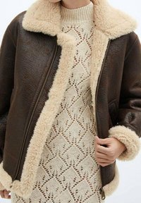 Veste en cuir marron avec col et poignets en shearling beige ; fermeture éclair ; portée sur une robe en tricot crème avec un motif texturé.