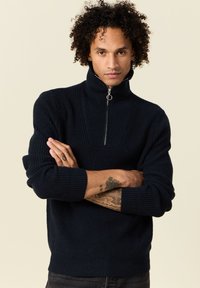 Pull en maille côtelée bleu foncé avec un col montant et une fermeture éclair mi-longue. Présente un tissu texturé et des poignets ajustés.