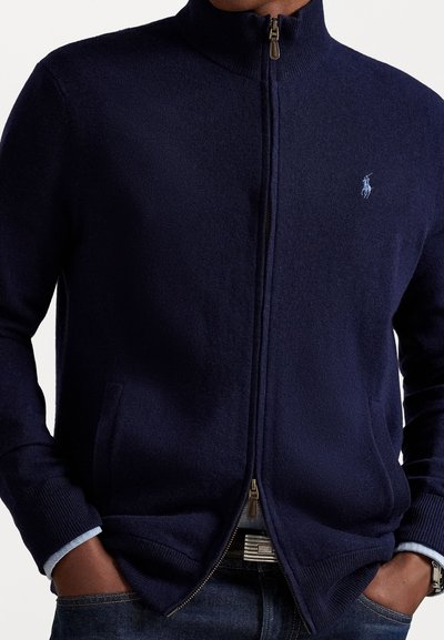 Granatowy sweter na zamek błyskawiczny z wysokim kołnierzem, prążkowanymi mankietami i bocznymi kieszeniami. Posiada małe haftowane logo na klatce piersiowej. Miękka textura.