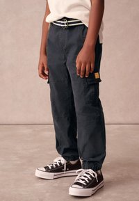 Mørke cargobukser med elastiske manchetter, udstyret med sidelommer og et lille orange mærke, kombineret med sorte lærreds-sneakers med hvide såler.