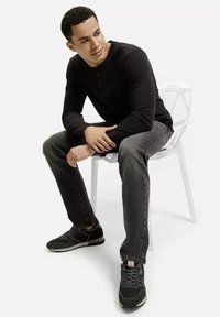 Mężczyzna siedzi na białym, geometrycznym krześle, ubrany w czarną, długosleeve koszulę henley, ciemnoszare dżinsy i czarne trampki.