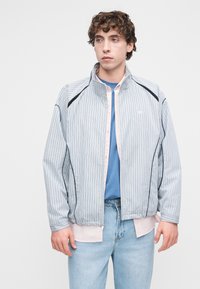 Μπουφάν με μπλε και λευκές ρίγες, φερμουάρ μπροστά, μαύρες λεπτομέρειες και ψηλό γιακά, φορεμένο πάνω από μπλε πουκάμισο και ανοιχτόχρωμο τζιν.