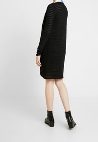 Černé pletené šaty s texturovaným vzorem, dlouhými rukávy, kulatým výstřihem a rovnou siluetou. Nosí se s černými kotníkovými botami.