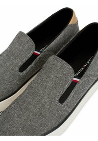 Gray slip-on shoes with fabric upper, elastic side panels, brown leather accents, and a white sole. Features branding with red and blue stripes.