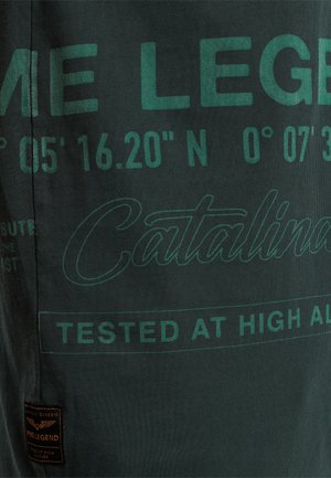 Tamnozeleni T-majica s ispisanim tekstom u svjetlijoj zelenoj boji. Detalji uključuju koordinate i riječ "Catalina" pisanom bojom, zajedno s oznakom od tkanine.