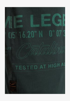 Tmavo zelené tričko s potlačeným textom v svetle zelenej. Detaily obsahujú súradnice a "Catalina" v písmovom písme, spolu s textilnou nálepkou.