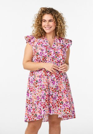 Usmievavá žena s kučeravými vlasmi, oblečená v bezrukávových šatách s kvetinovým vzorom s ružovými, červenými a fialovými kvetmi na bielom pozadí.