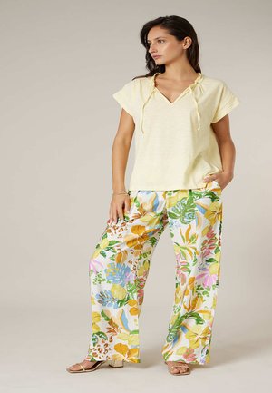 Femme debout portant un haut jaune pâle à manches courtes et un pantalon large à imprimé floral multicolore, posant avec une main dans la poche.
