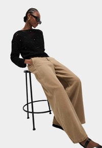 Pull en maille noir avec motif texturé, pantalon beige à jambes larges et lunettes de soleil noires, assis sur un tabouret noir devant un fond clair.