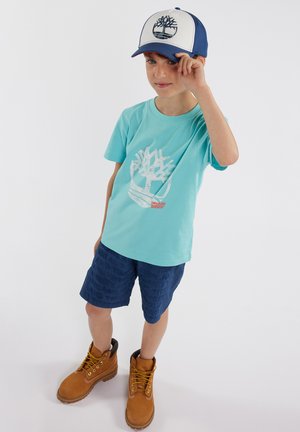 T-shirt en coton turquoise avec un graphique d'arbre, shorts à motifs navy, et bottes beige avec des lacets dorés, le tout complété par une casquette blanche et bleue.