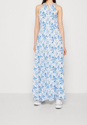 Kobieta w białej, bezrękawnikowej sukience maxi w niebieskie kwiaty, w białych platformowych trampkach, stojąca na jednolitym tle.