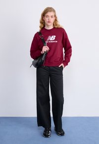 Sweatshirt bordeaux avec le logo "New Balance" blanc, associé à un pantalon large noir et des baskets noires, sur fond clair.